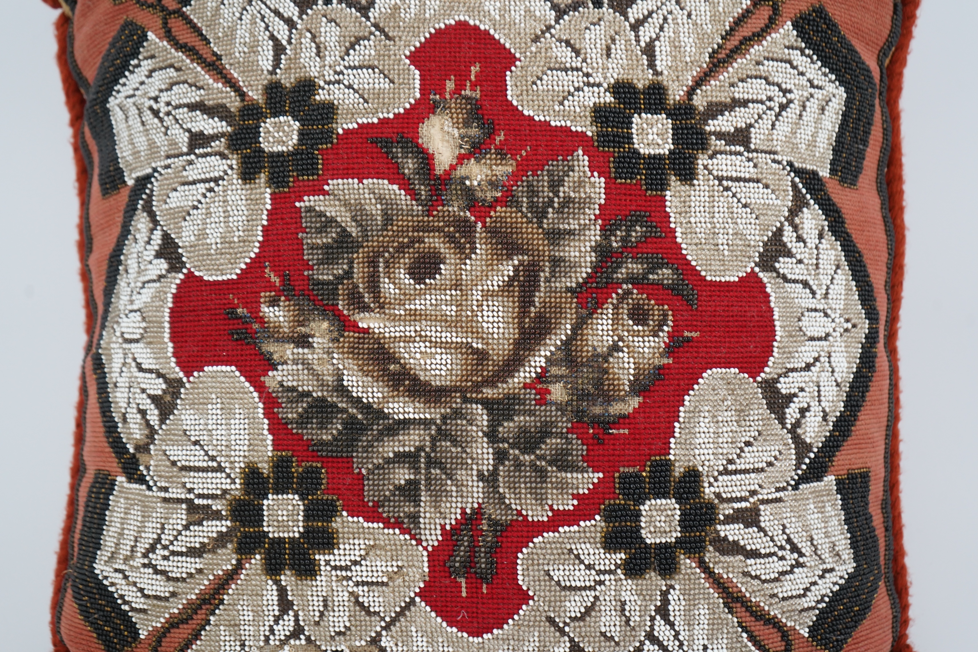 A 19th century Berlin bead worked cushion, the beadwork embroidered as a central rose, surrounded by an ornate tasseled swag, on a red wool worked background, 43cm x 45cm. Condition - some beadwork missing due to age and